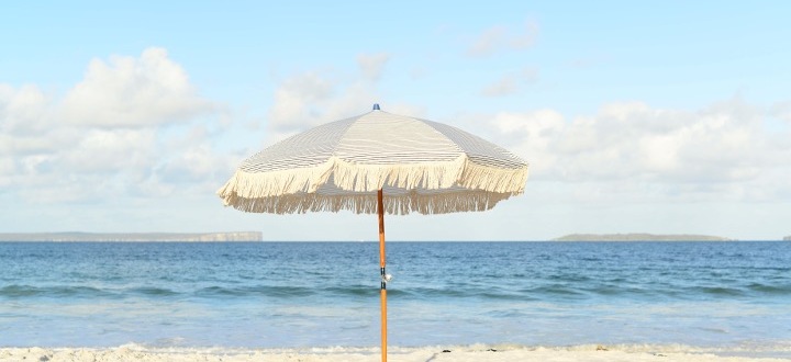 Ein beiger Strandparasol mit Fransen steht im Sand. Im Hintergrund erstreckt sich ein ruhiges, blaues Meer bis zum Horizont unter einem hellblauen Himmel mit wenigen Wolken. Das Bild vermittelt ein Gefühl von Ruhe und Sommer am Strand.