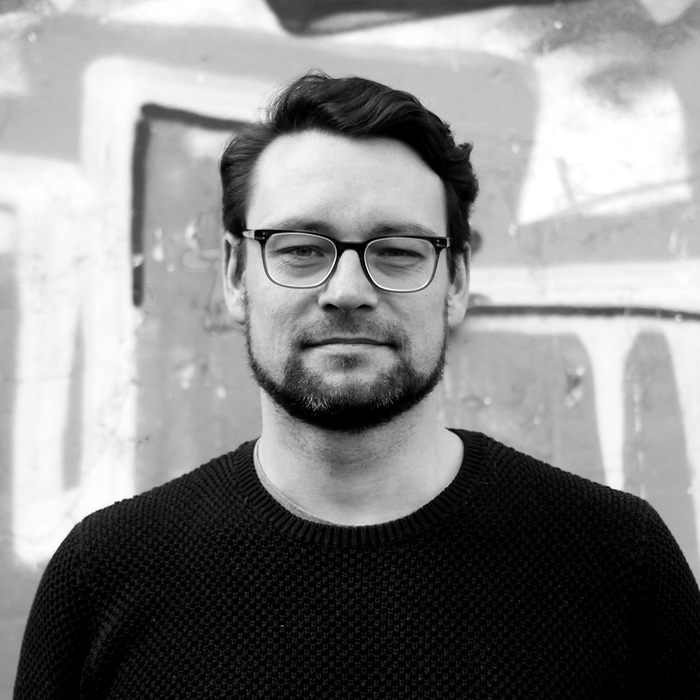 Schwarz-Weiß Porträt eines Mannes mit Brille. Er hat dunkles, lockiges Haar und einen Bart. Sein Blick ist freundlich und wirkt entspannt. Im Hintergrund ist eine unstrukturierte Wand zu sehen.