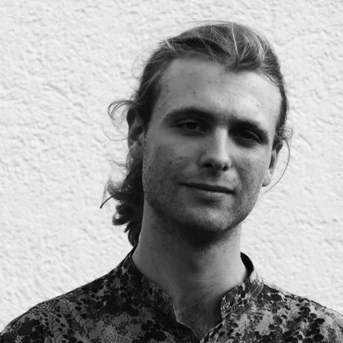 Schwarz-Weiß Porträt eines jungen Mannes mit langen, lockigen Haaren und Bart. Er blickt direkt in die Kamera und wirkt ruhig und gelassen. Er trägt ein Hemd mit einem floralen Muster. Der Hintergrund ist eine strukturierte, helle Wand.