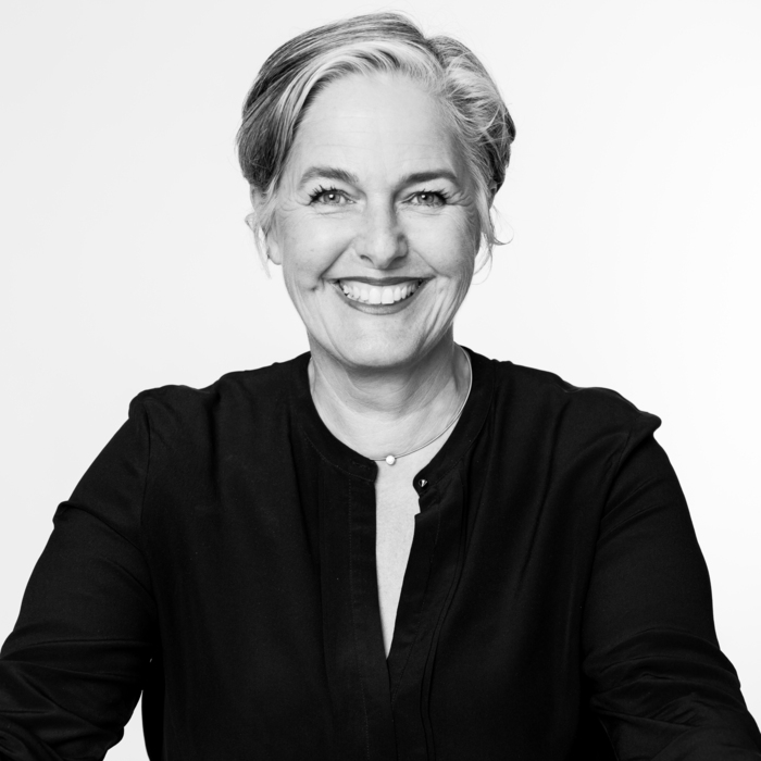 Schwarz-Weiß Porträt einer Frau mittleren Alters. Sie hat kurzes, helles Haar und ein freundliches Lächeln. Sie trägt ein dunkles Oberteil und wirkt sympathisch und offen.