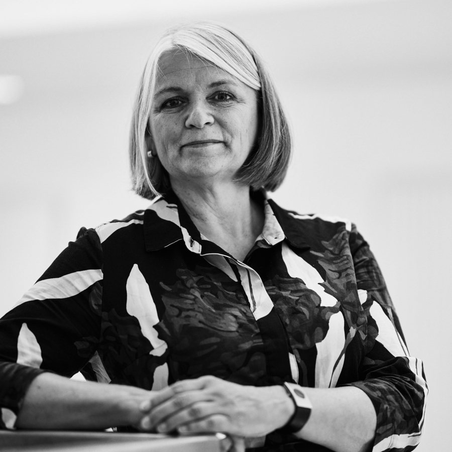 Schwarz-Weiß Porträt einer Frau mittleren Alters. Sie hat kurzes, helles Haar und trägt ein gemustertes Hemd. Ihr Blick ist direkt und wirkt ruhig und selbstbewusst. Sie sitzt an einem Tisch und hält ihre Hände gefaltet vor sich.