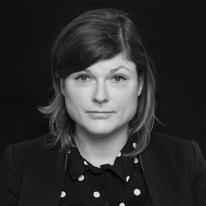 Schwarz-Weiß Porträt einer Frau. Sie hat mittellanges, gewelltes Haar und trägt ein Hemd mit weißen Punkten. Ihr Blick ist direkt und wirkt ruhig und selbstbewusst.