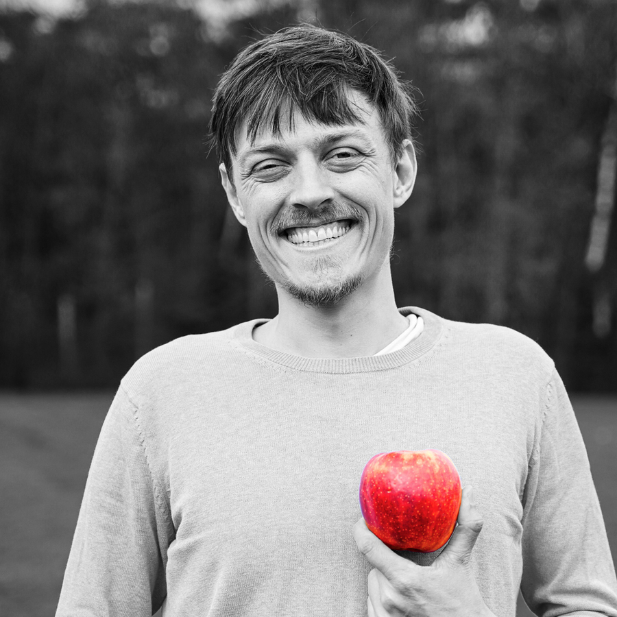 Ein lächelnder Mann hält einen roten Apfel in der Hand. Er hat kurze dunkle Haare und einen kurzen Bart. Er wirkt freundlich und zufrieden.