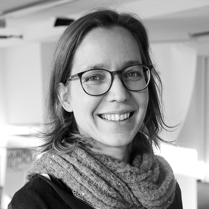 Porträt einer lächelnden Frau mit Brille und Schal. Sie wirkt freundlich und offen. Das Bild ist in Schwarz-Weiß gehalten.