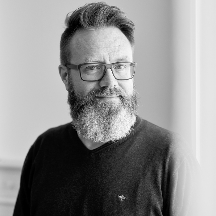 Schwarz-Weiß Porträt eines Mannes mit Vollbart. Er trägt eine Brille und einen dunklen Pullover. Sein Blick ist freundlich und wirkt einladend.