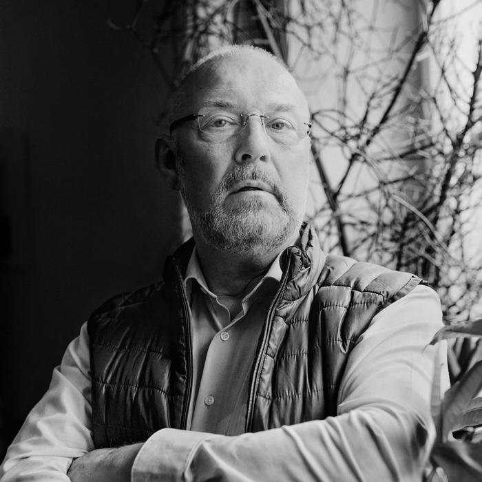 Schwarz-Weiß Porträt eines Mannes mittleren Alters mit Glatze und Vollbart. Er trägt eine Brille, ein Hemd und eine Weste. Sein Blick ist ernst und nachdenklich. Im Hintergrund sind Äste zu erkennen.