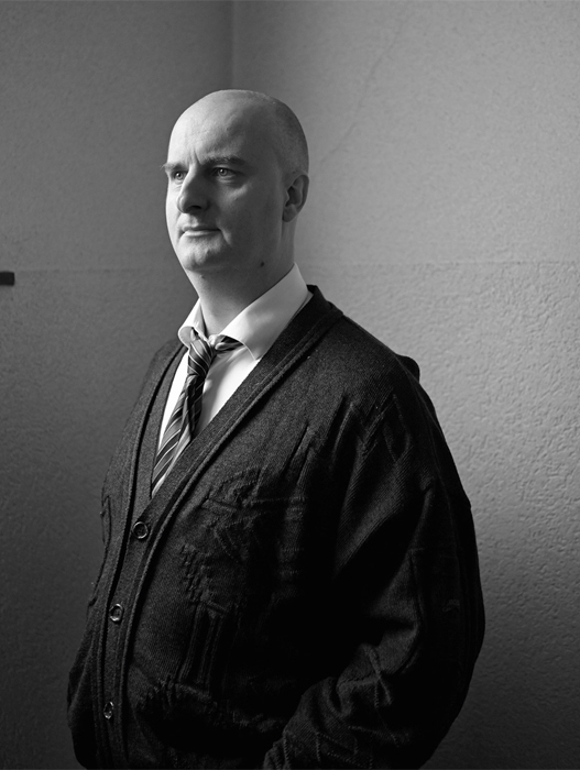 Schwarz-Weiß Porträt eines Mannes. Er ist glatzköpfig und trägt ein weißes Hemd mit einer Krawatte und einen karierten Strickcardigan. Er blickt nach rechts und wirkt nachdenklich.