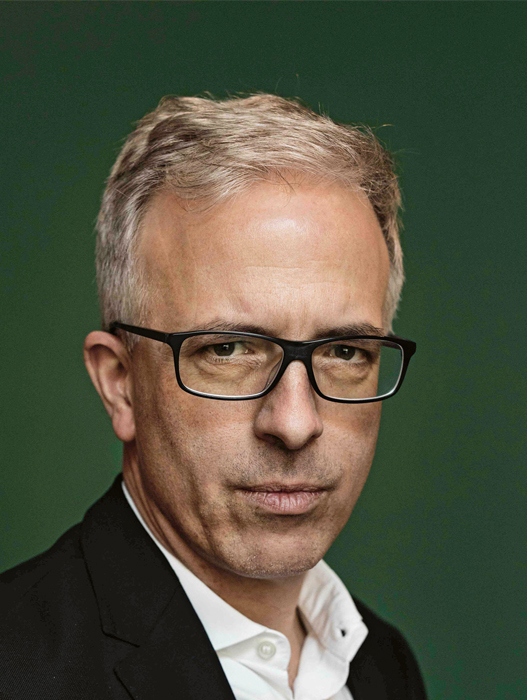 Porträt eines Mannes mittleren Alters mit grau meliertem Haar und Brille. Er trägt einen dunklen Anzug und ein weißes Hemd. Sein Blick ist direkt und wirkt konzentriert.