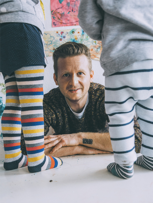 Ein lächelnder Mann sitzt auf dem Boden und schaut hoch zu zwei Kindern, die vor ihm stehen. Die Kinder sind nur von den Knien abwärts zu sehen, sie tragen bunte Socken. Im Hintergrund hängt ein farbenfrohes Bild an der Wand.