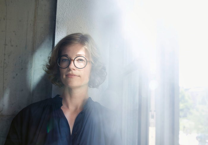 Eine Frau mit blonden, lockigen Haaren und runder Brille blickt ruhig in die Kamera. Sie trägt ein dunkles Hemd und steht vor einem Fenster, das von hellem Licht durchflutet wird. Ihr Gesichtsausdruck wirkt gelassen und nachdenklich.
