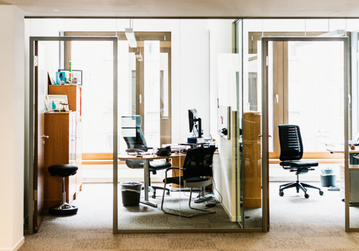 Ein helles Büro mit zwei Arbeitsplätzen, die durch Glaswände voneinander getrennt sind. In einem der Büros sitzt eine Person an einem Schreibtisch und arbeitet. Das Ambiente wirkt modern und aufgeräumt.
