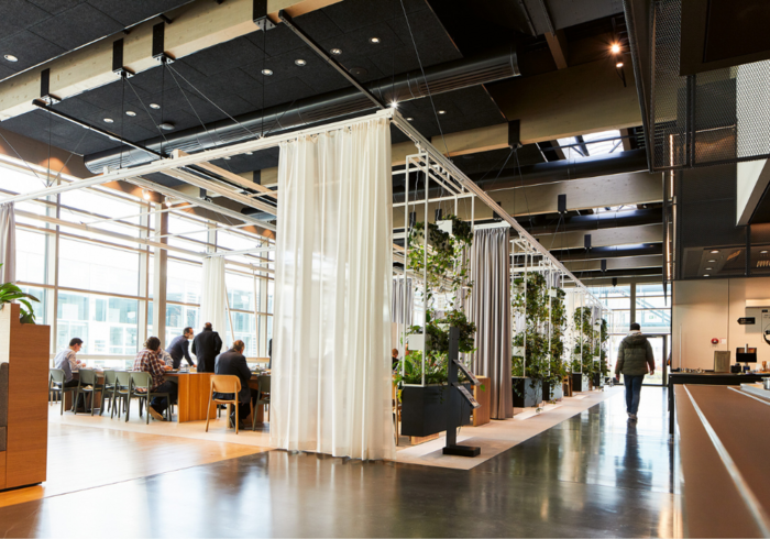Ein heller, offener Raum, vermutlich eine Großraumkantine oder ein Coworking-Space. Mehrere Personen sitzen an Tischen und unterhalten sich. Im Hintergrund eine große Fensterfront mit Tageslicht. Ein Mann geht im rechten Bildbereich entfernt in Richtung einer Theke. Der Raum ist modern eingerichtet mit viel Holz und Pflanzen, die als Raumteiler dienen.
