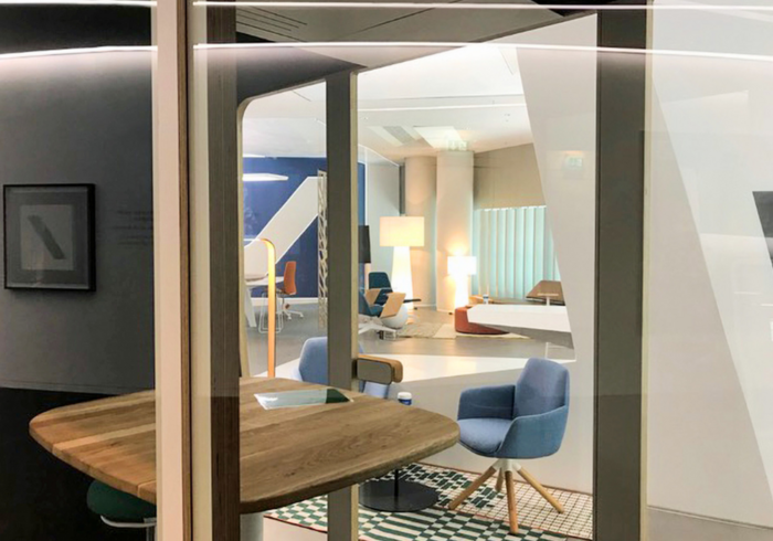 Ein moderner Büro- oder Empfangsbereich. Im Vordergrund eine halbrunde Holztheke. Durch einen Glastor ist ein heller Raum mit Schreibtischen, Stühlen und Stehlampen zu sehen. Das Ambiente wirkt offen und einladend.