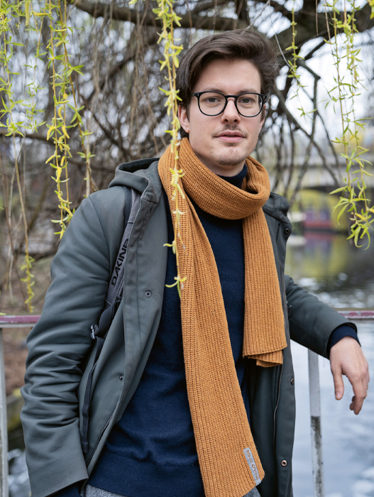 Ein junger Mann mit dunklen Haaren und Brille blickt selbstbewusst in die Kamera. Er trägt eine grüne Jacke, einen dunklen Pullover und einen orangefarbenen Schal. Im Hintergrund sind Bäume mit jungen Blättern zu sehen.