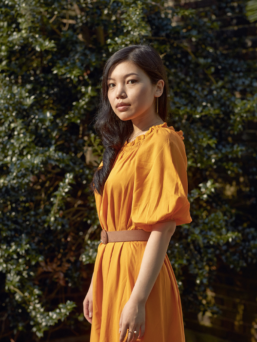 Eine junge Frau mit langen, dunklen Haaren steht vor einem grünen Hintergrund aus Blättern. Sie trägt ein oranges Kleid mit kurzen Ärmeln und einen schmalen Gürtel. Ihr Blick ist ruhig und sie wirkt selbstbewusst.