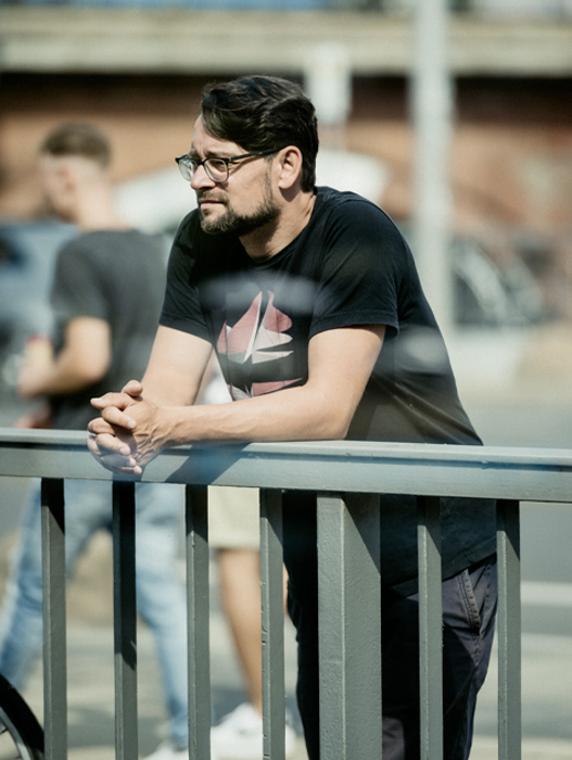 Ein Mann mit dunklem Haar und Bart lehnt nachdenklich an einer Metallreling. Er trägt ein schwarzes T-Shirt mit einem weißen Aufdruck und dunkle Jeans. Im Hintergrund sind verschwommene Personen zu erkennen, die auf einer Straße stehen. Das Bild vermittelt eine ruhige, introspektive Stimmung.