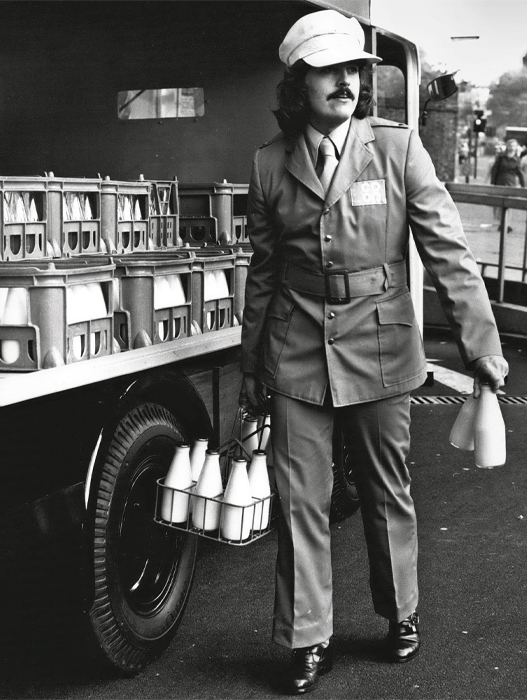 Schwarz-Weißfoto eines Milchmanns in Uniform. Er trägt eine weiße Kappe, einen braunen Anzug mit Gürtel und eine Krawatte. Er hält einen Korb mit Milchflaschen in der Hand und trägt weitere Flaschen unter dem Arm. Im Hintergrund ist ein Lieferwagen voller Milchflaschen zu sehen. Der Mann wirkt professionell und konzentriert.