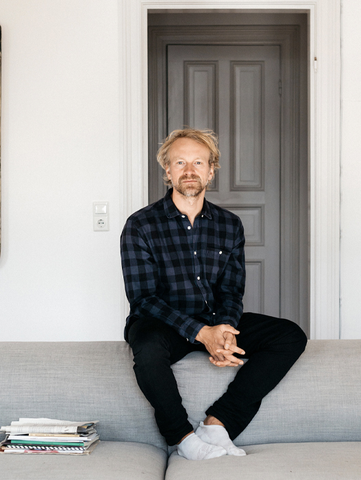 Ein Mann sitzt entspannt auf einer hellgrauen Couch in einem minimalistisch eingerichteten Raum. Er trägt ein kariertes Hemd und dunkle Hosen. Sein Blick ist ruhig und nachdenklich. Im Hintergrund befindet sich eine Tür in einem hellen Rahmen.