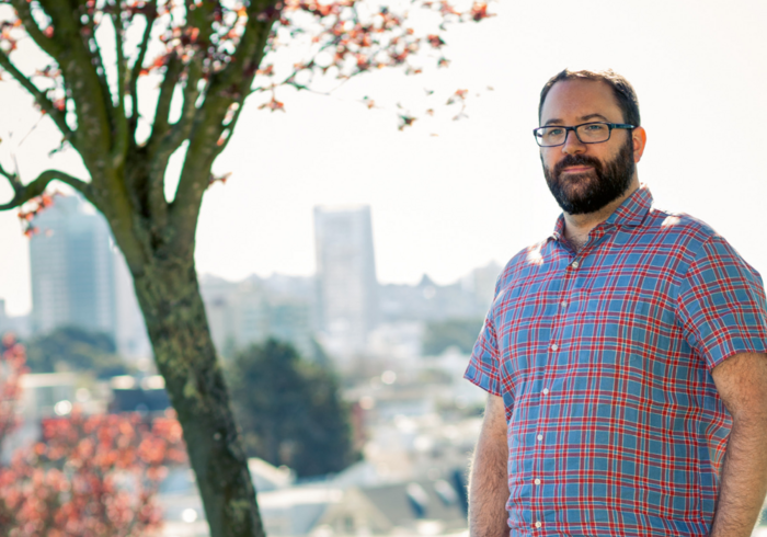 Ein Mann mit Bart und Brille steht lächelnd im Vordergrund. Er trägt ein blau-kariertes Hemd. Im Hintergrund ist eine Stadtlandschaft mit Hochhäusern und ein Baum mit roten Blüten zu sehen. Das Bild vermittelt einen freundlichen und entspannten Eindruck.