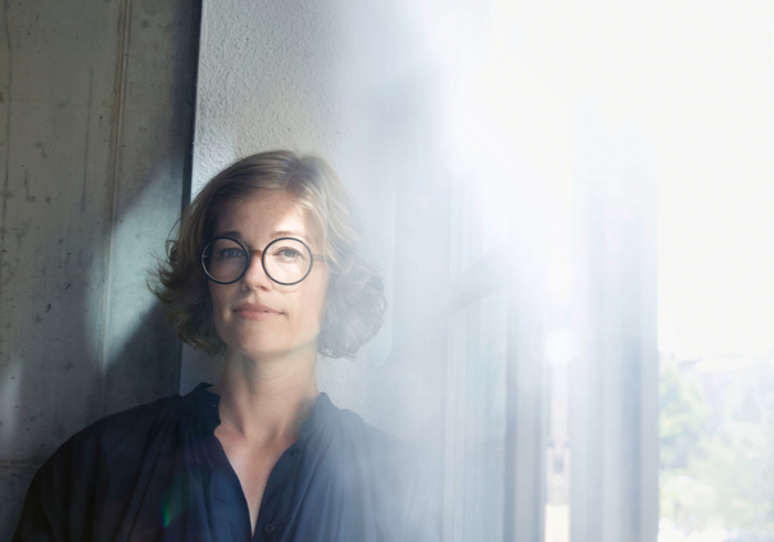 Eine Frau mit blonden, kurzen Haaren und runder Brille blickt nachdenklich aus einem Fenster. Das Licht scheint hell hinter ihr und erzeugt einen weichen, verschwommenen Hintergrund. Sie trägt ein dunkles Hemd und wirkt ruhig und introspektiv.