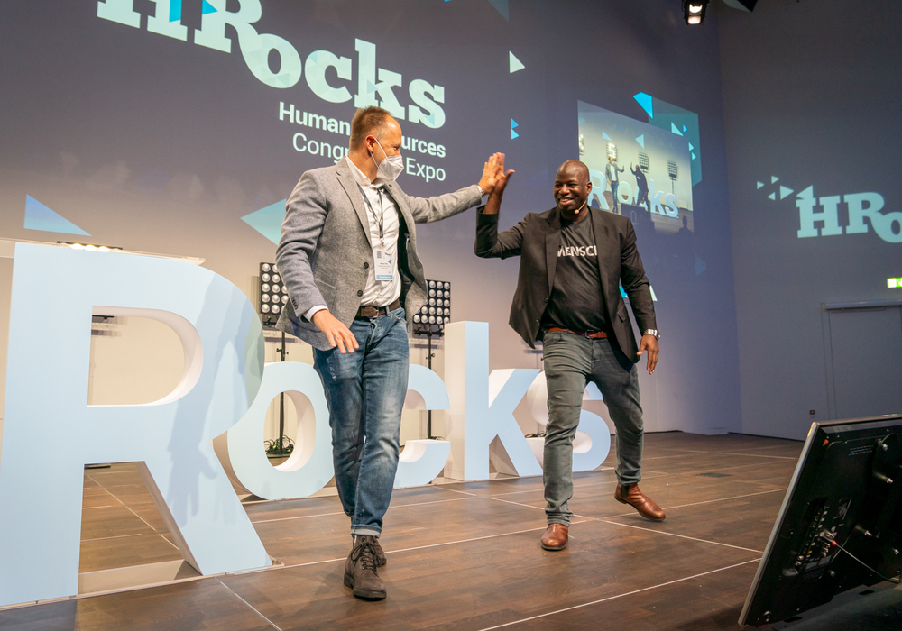 Zwei Männer stehen auf einer Bühne vor einem großen Banner mit der Aufschrift "HR Rocks". Der Mann links trägt einen beigen Blazer und Jeans, er lächelt und hebt die rechte Hand zum High-Five. Der Mann rechts, mit dunkler Hautfarbe, trägt ein schwarzes Hemd und rote Hosen, er erwidert den High-Five. Im Hintergrund ist eine Projektion mit Gebäuden zu sehen. Die Atmosphäre wirkt positiv und dynamisch, wie nach einem erfolgreichen Auftritt oder einer Präsentation.