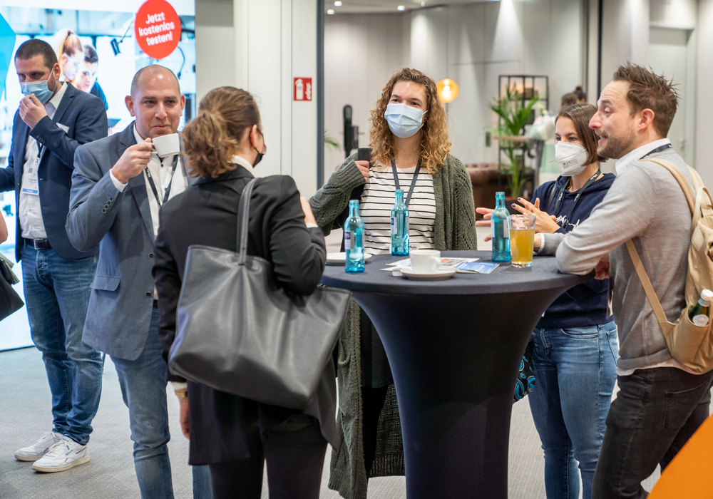 Das Bild zeigt eine Gruppe von Menschen, die um einen Tisch versammelt sind und sich unterhalten. Einige Personen tragen Masken, was auf ein mögliches Gesundheits- oder Sicherheitsrisiko hinweist. Der Tisch ist mit verschiedenen Gegenständen bedeckt, darunter Flaschen, Tassen und eine Schüssel. Die Menschen stehen sich gegenüber und vermitteln so den Eindruck von Interaktion und Engagement. Im Hintergrund ist ein Schild mit der Aufschrift "Jetzt konserviert" zu sehen, was darauf hindeutet, dass es sich um einen professionellen oder geschäftlichen Ort handeln könnte.