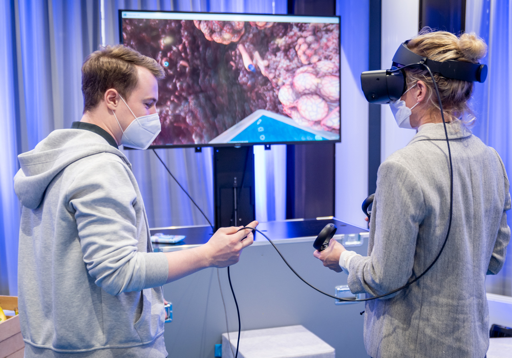 Zwei Personen mit Virtual-Reality-Headsets stehen vor einem großen Bildschirm, auf dem das Bild eines Korallenriffs zu sehen ist. Die Person auf der linken Seite hält eine Fernbedienung in der Hand, während die Person auf der rechten Seite ein Telefon in der Hand hält.