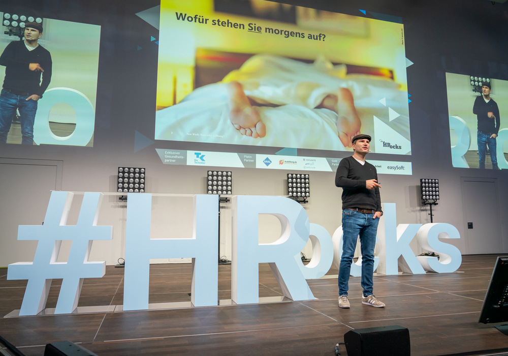 Ein Mann in Jeans und Jacke steht auf einer Bühne und hält einen Vortrag. Im Hintergrund eine große Leinwand mit der Frage "Wofür stehen Sie morgens auf?". Unterhalb der Leinwand befindet sich ein großes, hellblaues Hashtag "#HRrocks" mit den Logos verschiedener Unternehmen. Das Publikum ist im Hintergrund angedeutet.