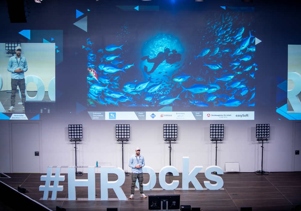 Das Bild zeigt eine Person, die auf einer Bühne steht, hinter der ein großer Bildschirm mit einer Unterwasserszene mit Fischen zu sehen ist. Die Bühne ist für eine Präsentation oder eine Veranstaltung hergerichtet, und um die Bühne herum sind mehrere Lichter und Lautsprecher angeordnet. Das Bild enthält auch den Hashtag "#HRrocks", der deutlich im Vordergrund zu sehen ist.