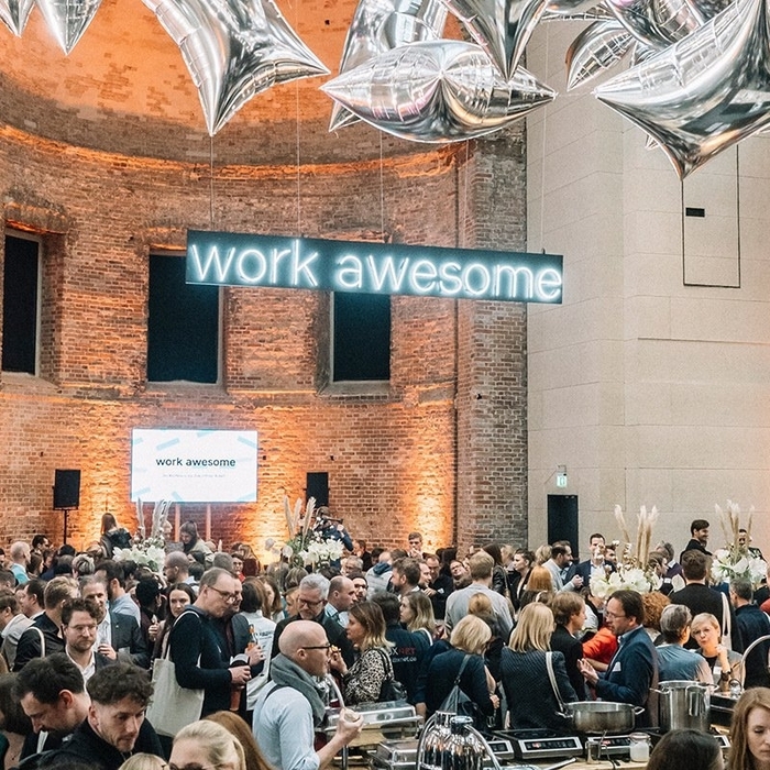 Große Menschenmenge in einem hellen Raum mit Backsteinwänden. Im Hintergrund hängt ein großes Schild mit der Aufschrift "work awesome". Menschen stehen und unterhalten sich, einige scheinen an einem Buffet zu stehen. Die Atmosphäre wirkt festlich und gesellig.