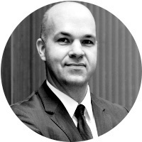 Schwarz-Weiß Porträt eines Mannes mit Glatze und dunklem Anzug. Er lächelt freundlich und wirkt sympathisch. Im Hintergrund ist eine verschwommene Struktur zu erkennen.