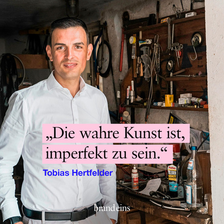 Ein Mann mittleren Alters mit dunklem Haar und einem gepflegten Bart lächelt in die Kamera. Er trägt ein weißes Hemd und einen dunklen Gürtel. Im Hintergrund befindet sich eine Werkstatt mit Werkzeugen und Regalen. Ein pinkfarbener Kasten mit dem Zitat „Die wahre Kunst ist, perfekt zu sein.“ und dem Namen „Tobias Hertfelder“ überlagert das Bild.