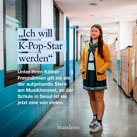Ein junges Mädchen steht selbstbewusst in einem hellen Flur einer Schule. Sie hat lange, dunkle Haare und trägt eine Schuluniform mit Krawatte. Sie blickt in die Ferne, wirkt entschlossen und hoffnungsvoll. Im Hintergrund sind Schließfächer und ein Fenster zu sehen. Der Text auf dem Bild deutet darauf hin, dass sie davon träumt, ein K-Pop-Star zu werden.