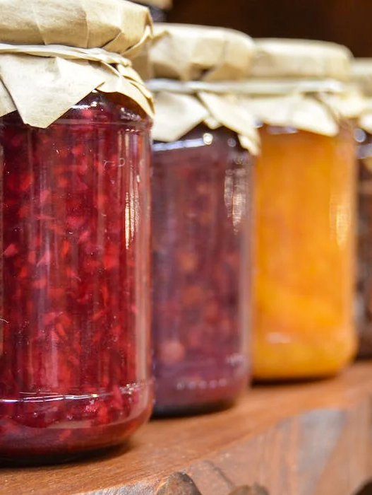 Drei Gläser selbstgemachter Marmelade stehen nebeneinander auf einem Holzbrett. Das linke Glas enthält rote Beerenmarmelade, das mittlere dunkle Kirschmarmelade und das rechte orangefarbene Aprikosenmarmelade. Alle Gläser sind mit braunem Papier verschlossen.