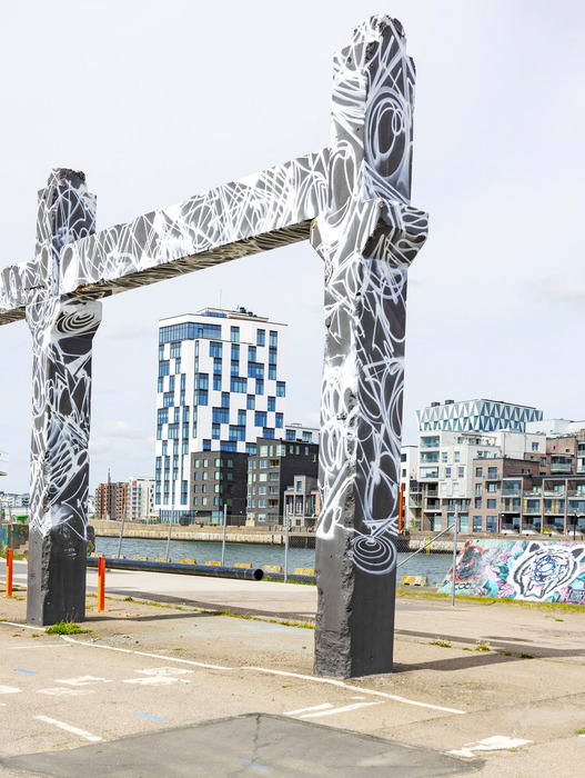 Ein großes, modernes Tor-ähnliches Kunstwerk aus Beton steht auf einem Parkplatz am Wasser. Das Tor ist mit einem komplexen Muster aus schwarzen und weißen Linien bemalt, das an organische Formen oder Wellen erinnert. Im Hintergrund erstrecken sich moderne Gebäude mit vielen Fenstern und einem hellen Farbton. Das Wasser glänzt leicht, und am Horizont sind weitere Gebäude und ein blauer Himmel zu sehen.