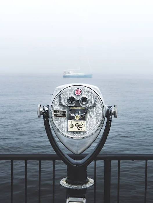 Ein metallener Fernrohr-Automat steht auf einer Geländer vor dem Meer. Im Hintergrund ist ein Schiff im Dunst zu erkennen. Das Ambiente ist ruhig und neblig.