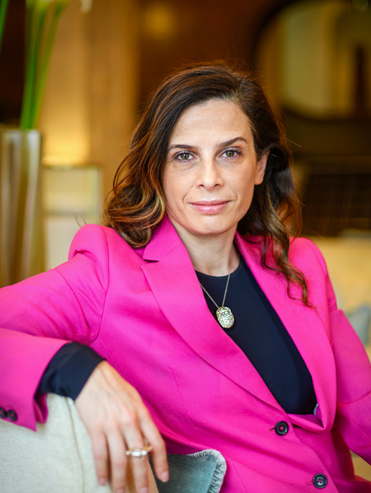 Eine Frau mit braunen Haaren sitzt entspannt in einem Sessel. Sie trägt einen auffälligen pinkfarbenen Blazer und ein dunkles Oberteil. Sie lächelt leicht und wirkt freundlich und zugewandt. Im Hintergrund ist ein heller Raum mit Pflanzen zu erkennen.