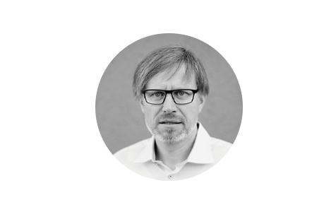 Schwarz-Weiß Porträt eines Mannes mittleren Alters mit kurzgeschnittenem, grau meliertem Haar und Brille. Er blickt direkt in die Kamera, sein Gesichtsausdruck wirkt ernst und konzentriert.