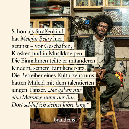 Ein Mann mittleren Alters mit dunklem Haar und Bart sitzt lächelnd in einem Raum. Er trägt ein kariertes Hemd und hält ein Mikrofon in der Hand. Im Hintergrund sind Regale mit Büchern und Schallplatten zu sehen. Der Text im Bild erzählt, dass der Mann als Straßenkind tanzte und später von einem Kulturzentrum unterstützt wurde.