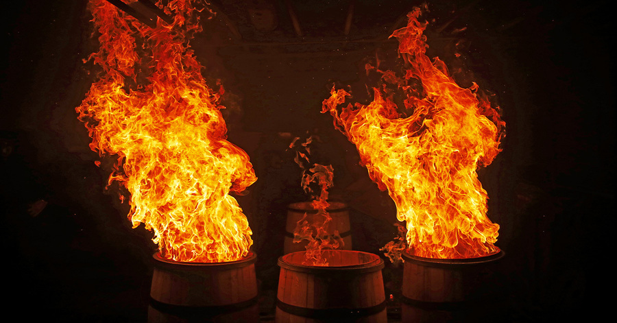 Zwei große Fässer stehen im Dunkeln und sind von hohen Flammen umhüllt. Die Flammen leuchten in warmen Orange- und Gelbtönen und erzeugen eine dramatische Atmosphäre. Im Hintergrund sind schemenhaft weitere Objekte und Personen erkennbar, die jedoch nicht klar zu identifizieren sind.