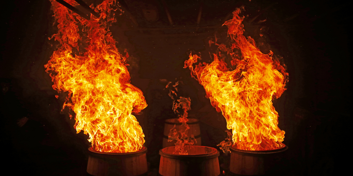 Zwei brennende Fässer stehen in einem dunklen Raum. Die Flammen schlagen hoch und erhellen die Umgebung. Im Hintergrund sind weitere Fässer zu erkennen.