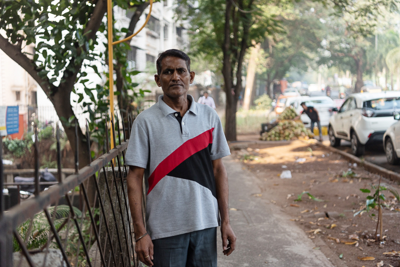 Ein älterer Mann steht auf einem Gehweg in einer Stadt. Er trägt ein graues Polohemd mit roten und schwarzen Streifen sowie Jeans. Er wirkt nachdenklich und blickt geradeaus. Im Hintergrund sind Gebäude, Bäume und parkende Autos zu sehen.