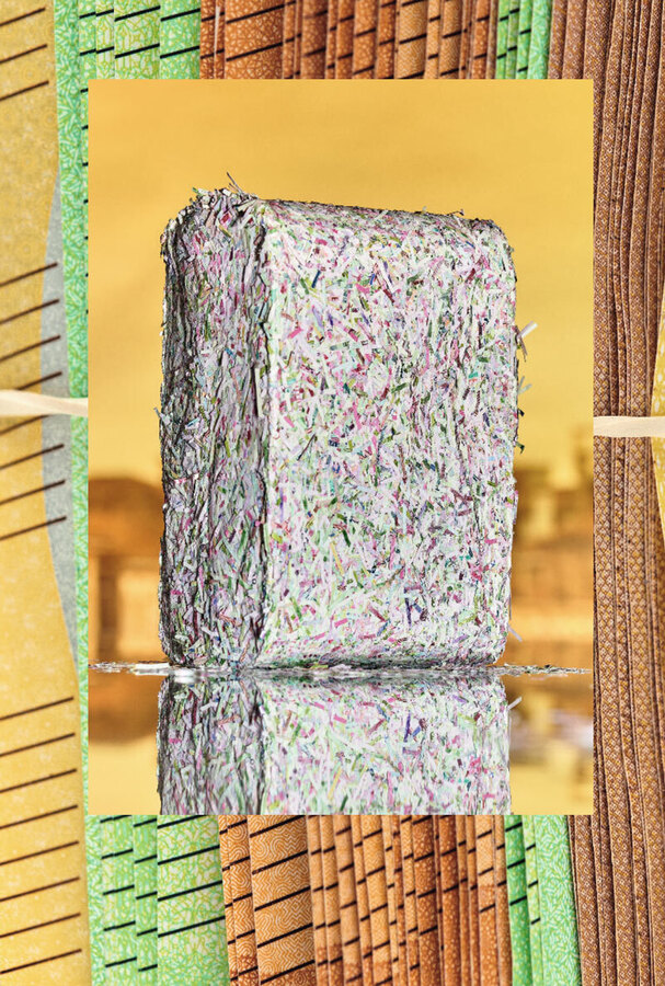 Ein quaderförmiger Ballen aus buntem Papierabfall steht auf einer spiegelnden Oberfläche. Das Papier besteht aus vielen kleinen, farbigen Fragmenten. Der Hintergrund ist ein abstraktes Muster aus vertikalen Linien in verschiedenen Farben.