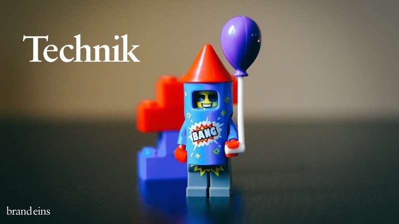 Ein Lego-Minifigur, gekleidet als Rakete, hält einen blauen Ballon. Im Hintergrund befindet sich ein rotes Lego-Gebäude. Das Bild vermittelt eine spielerische und feierliche Atmosphäre, möglicherweise zu einem Geburtstag oder einer anderen festlichen Gelegenheit.