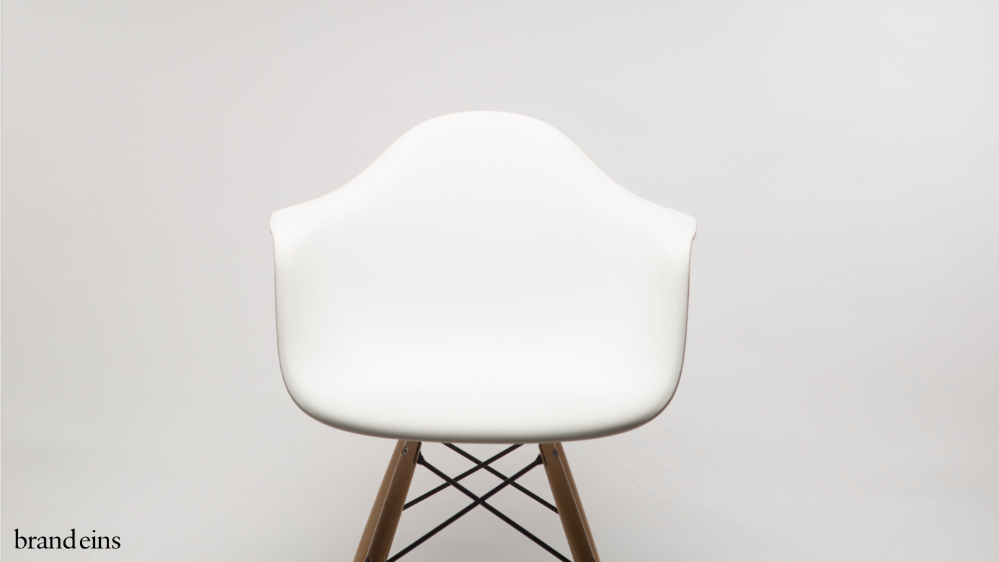 Ein weißer Eames-Sessel steht vor einem hellgrauen Hintergrund. Der Sessel hat eine geschwungene Form und steht auf schlanken, hellen Holzbeinen.
