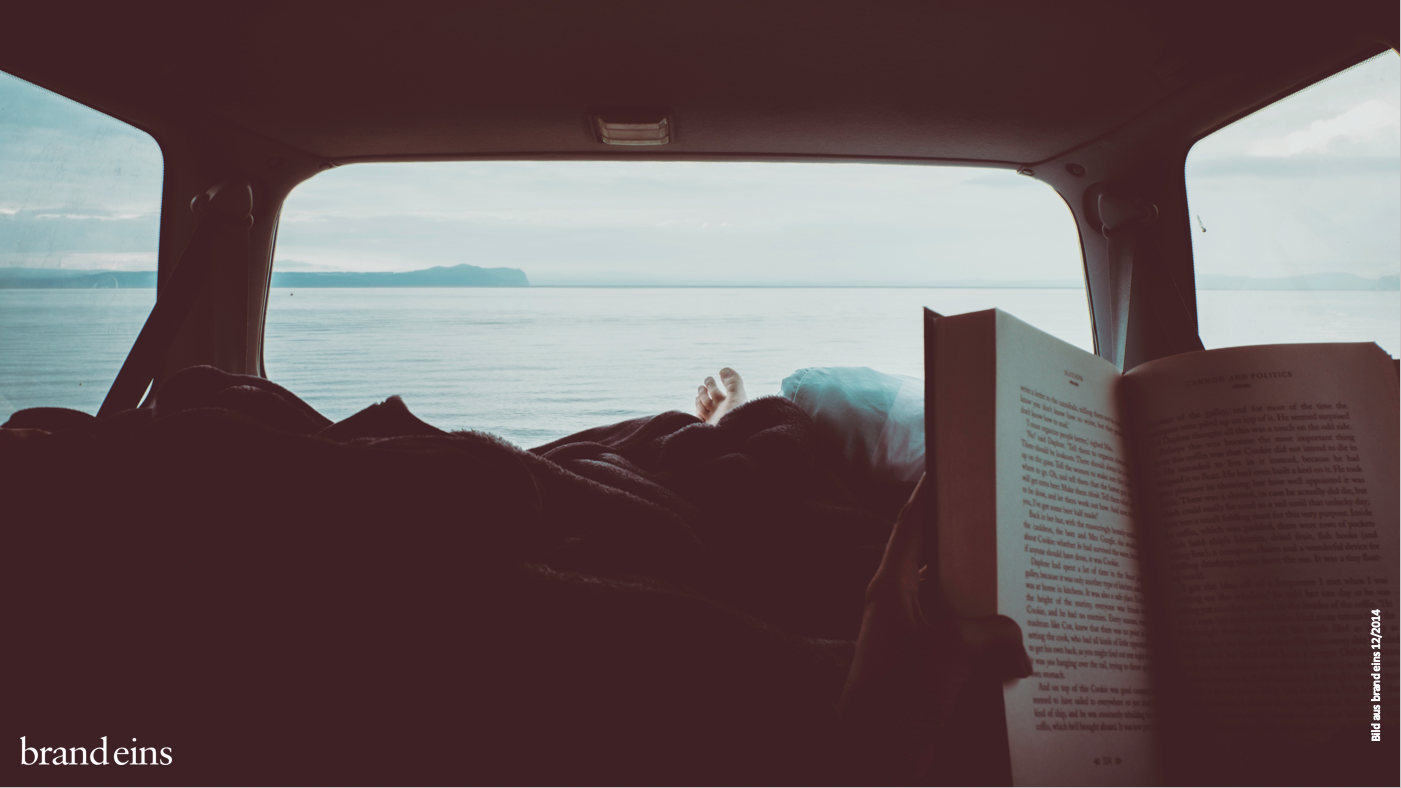 Ein Mann liegt entspannt im Kofferraum eines Autos und liest ein Buch. Er blickt auf das Meer hinaus, das durch die geöffnete Heckklappe sichtbar ist. Die Szene wirkt ruhig und friedlich, er scheint die Zeit zu genießen.