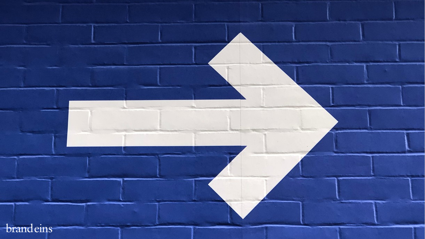 Ein weißer Pfeil, der nach rechts zeigt, ist auf einer dunklen Ziegelmauer gemalt. Der Pfeil symbolisiert eine Richtung oder einen Wegweiser.