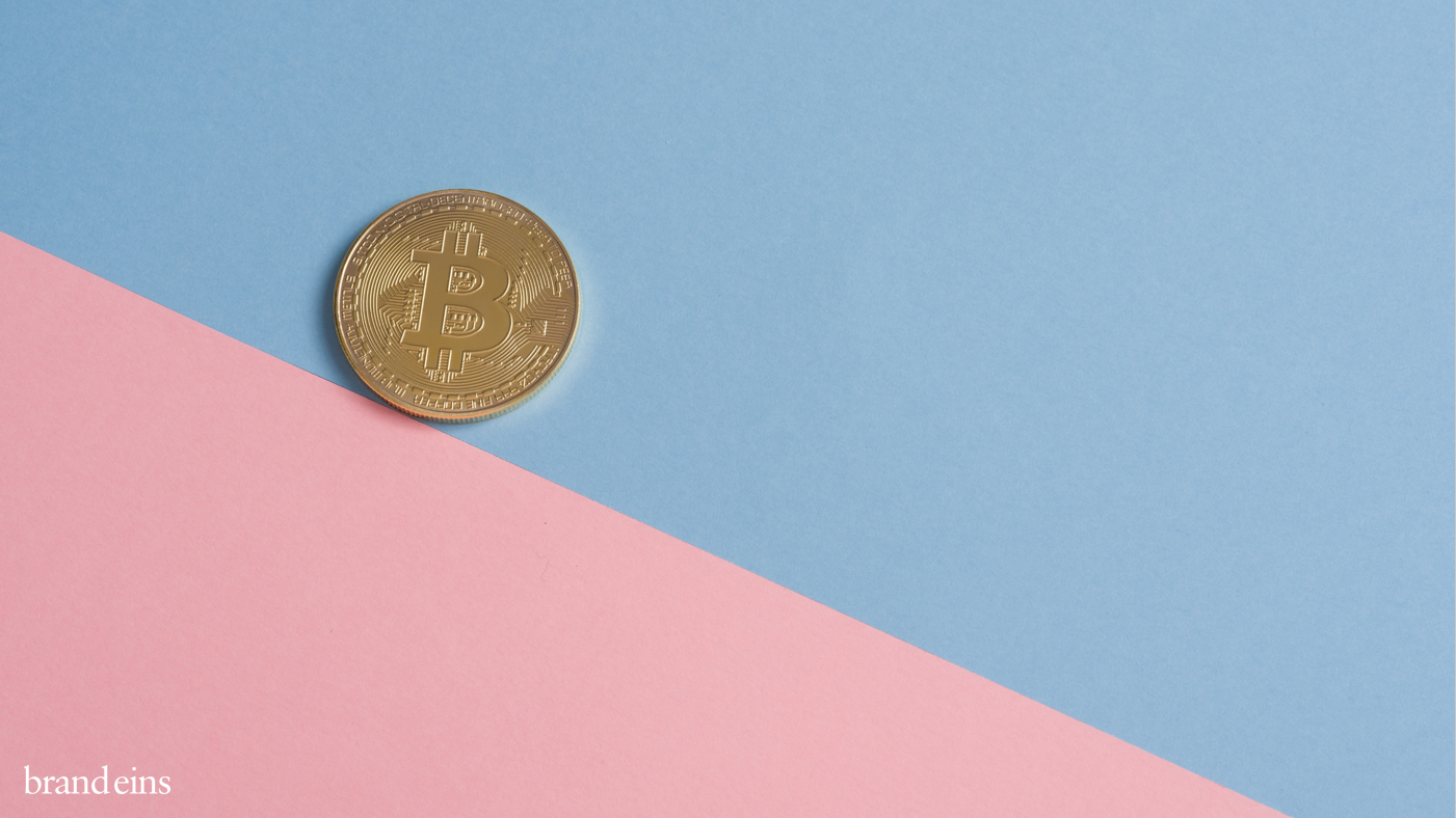 Ein goldfarbenes Bitcoin-Symbol liegt auf einem Hintergrund, der diagonal in hellblau und zartrosa geteilt ist. Der schwarze Bereich befindet sich unten rechts im Bild.