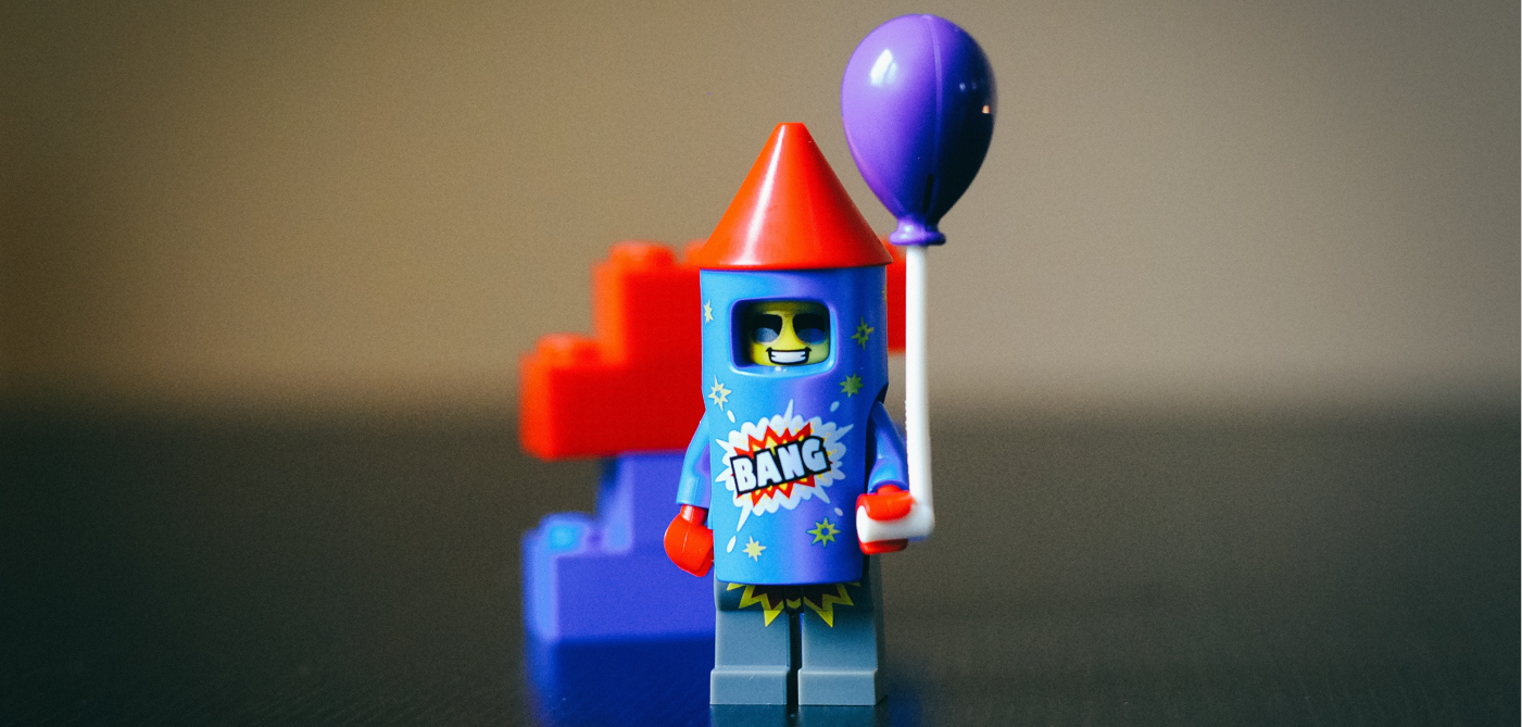 Ein LEGO-Minifigur als Rakete in Rot steht auf einer dunklen Oberfläche. Die Figur hält einen lila Ballon und wirkt fröhlich. Im Hintergrund sind unscharfe LEGO-Steine zu erkennen.