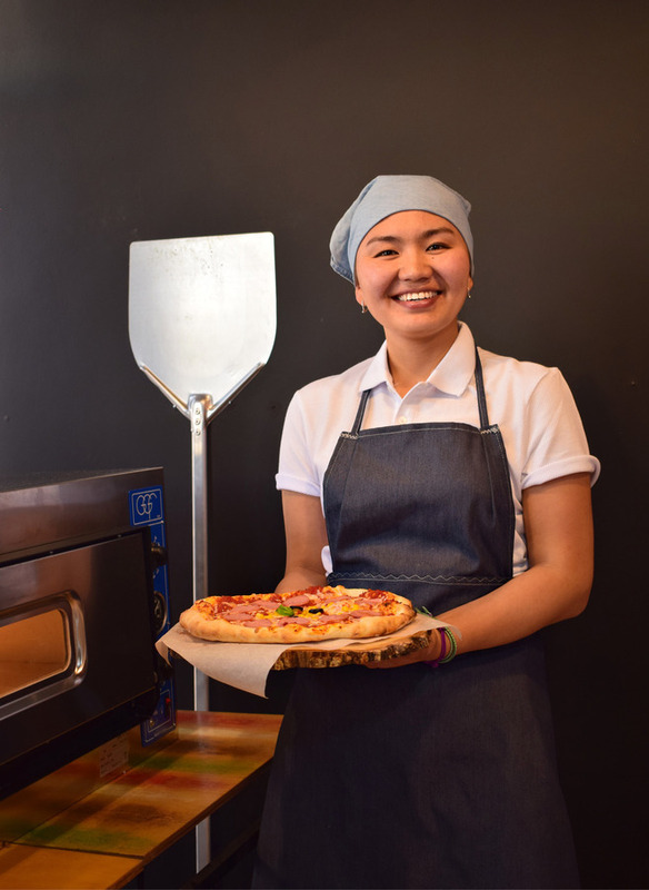 Eine junge Frau mit dunklen Haaren und freundlichem Lächeln hält eine frisch gebackene Pizza vor einem Ofen. Sie trägt eine blaue Kochhaube und eine dunkle Schürze und wirkt stolz auf ihre Arbeit. Im Hintergrund ist ein Pizzaofen und ein Pizzaschieber zu sehen.